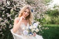 An attractive bride with a bouquet of wild flowers is sitting in a chair in a blooming garden