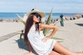 Attractive brunette girl with long hair is lying on the beach near sea. She wears hat, sunglasses and white short dress Royalty Free Stock Photo