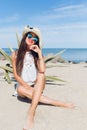 Attractive brunette girl with long hair is sitting on the beach near sea. She wears hat, sunglasses and white short Royalty Free Stock Photo