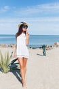 Attractive brunette girl with long hair is walking on the beach near sea. She wears hat, sunglasses and white short Royalty Free Stock Photo