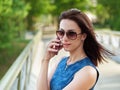 Attractive brunette woman in sunglasses and blue jeans dress has emotional phone conversation on mobile phone on park bridge outdo Royalty Free Stock Photo