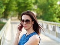 Attractive brunette woman in sunglasses and blue jeans dress has emotional phone conversation on mobile phone on park bridge outdo Royalty Free Stock Photo