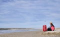 Attractive brunette travel girl sitting with a red suitcase by the sea, against the blue sky, place for text Royalty Free Stock Photo