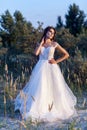 Attractive brunette woman with makeup and hairstyle wearing white wedding dress while posing at glade. standing with hand on waist Royalty Free Stock Photo