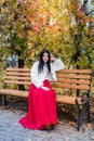 Attractive brunette woman in red dress sitting on bench in autumn park Royalty Free Stock Photo