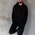 Attractive brutal young man in a fashionable black sweatshirt in vintage black jeans with a stylish beard posing on the street