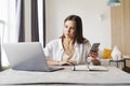 Attractive business woman sits at table in front of laptop and uses smartphone for work.