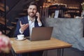 Attractive young elegant businessman talking on phone at laptop in cafe and smiling. Conversation.