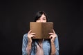 Attractive caucasian brunette girl in a shirt covering her face with a book and reading isolated on a black studio background. Royalty Free Stock Photo