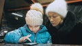 Attractive caucasian mother and daughter using smartphone sitting in cafe. Advanced little child shows and explains to Royalty Free Stock Photo