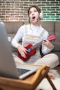 An attractive Caucasian young woman in headphones with a ukulele in her hands sings expressively at home next to a sofa