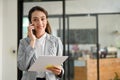 Attractive Asian businesswoman talking on the phone, holding a document, standing in her office Royalty Free Stock Photo