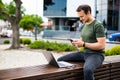 Attractive confident young man sitting on a bench outdoors at the city street, working on laptop computer, using mobile phone Royalty Free Stock Photo