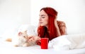 Attractive, contented, young, red-haired woman lying relaxed in bed with her cat with her coffee in a red cup, copy space Royalty Free Stock Photo