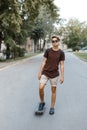 Attractive cool young hipster man in sunglasses in a stylish T-shirt in summer shorts in sneakers riding a skateboard in the park