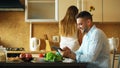 Attractive couple chatting in the kitchen early morning. Handsome man using tablet while his girlfriend cooking Royalty Free Stock Photo