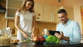 Attractive couple chatting in the kitchen early morning. Handsome man using tablet while his girlfriend cooking Royalty Free Stock Photo