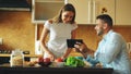 Attractive couple chatting in the kitchen early morning. Handsome man using tablet while his girlfriend cooking Royalty Free Stock Photo