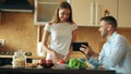 Attractive couple chatting in the kitchen early morning. Handsome man using tablet while his girlfriend cooking Royalty Free Stock Photo