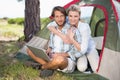 Attractive couple sitting by their tent using laptop smiling at camera