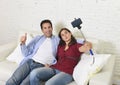 Attractive couple taking selfie photo or shooting self video with mobile phone and stick sitting at home couch smiling happy Royalty Free Stock Photo