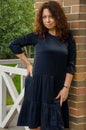 Attractive curly-haired red-haired woman for forty years standing on the porch of a country house in a blue dress with ruches. Royalty Free Stock Photo