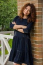 Attractive curly-haired red-haired woman for forty years standing on the porch of a country house in a blue dress with ruches. Royalty Free Stock Photo