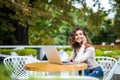 Attractive curly latin female freelancer hold smart phone while sitting at wooden table front open computer in modern coffee shop. Royalty Free Stock Photo