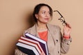 Attractive middle-aged female teacher holding eyeglasses, looking away, posing with stack of books, isolated background Royalty Free Stock Photo