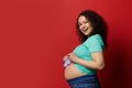 Attractive smiling ethnic pregnant woman with bare belly, posing with cute blue knitted baby booties, red background.