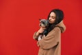 Attractive dog-owner female is posing on an isolated red background, holding her cute friend - yorkshire terrier