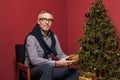 Attractive elderly man holds Christmas presents gold box against red studio wall background and green decorated Christmas tree Royalty Free Stock Photo