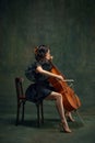 Attractive, elegant young woman, cellist, musician in black dress sitting on chair and playing cello against dark Royalty Free Stock Photo