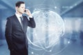 Attractive european businessman with global interface hologram talking on the phone and standing in blurry office interior. Royalty Free Stock Photo