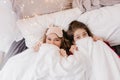 Attractive fair-haired girl in pink sleepmask hiding under blanket. Indoor photo of two refined sisters joking during