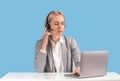 Attractive female helpline operator with headset and laptop answering client`s call at table against blue background Royalty Free Stock Photo