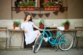 Attractive female in white dress and straw hat sitting outside at cosy cafe zone next to her blue vintage bike Royalty Free Stock Photo