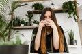 Attractive formal lady is sitting in stylish cafe with greenery and drinking coffee from a cup, looking into the camera. Stylish