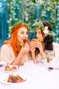 Attractive funny and hungry young bride and her bridesmaid in green floral wreath on heads eating hamburgers, while Royalty Free Stock Photo