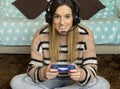 Attractive gamer girl with headphones plays with a wireless gamepad while looking at the screen in front of her. Young blonde girl