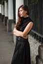 Attractive girl in a black dress with a corset on a background of a fence with clones. Evening walk on the city street.