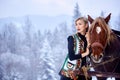 Attractive girl in embroidered sleeveless sheepskin fur coat at pair of strong horses in harness. Royalty Free Stock Photo