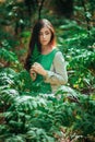 Attractive girl in a green medieval dress sits in the bushes of fern. Village woman herbalist collects herbs for potion in the for