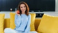 Attractive girl holds fresh red apple while sitting on yellow sofa at the stylish kitchen. Royalty Free Stock Photo