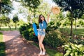 Attractive girl with long curly hair is posing with skateboard in summer park. She wears jeans shorts, jerkin