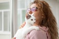 Attractive girl in pink clothes and eyeglasses with withe cat in her arms standing near the window at home. Pet owner Royalty Free Stock Photo