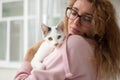 Attractive girl in pink clothes and eyeglasses with withe cat in her arms standing near the window at home. Pet owner Royalty Free Stock Photo