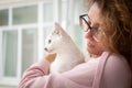 Attractive girl in pink clothes and eyeglasses with withe cat in her arms standing near the window at home. Pet owner Royalty Free Stock Photo