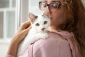 Attractive girl in pink clothes and eyeglasses with withe cat in her arms standing near the window at home. Pet owner Royalty Free Stock Photo