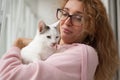 Attractive girl in pink clothes and eyeglasses with withe cat in her arms standing near the window at home. Pet owner Royalty Free Stock Photo